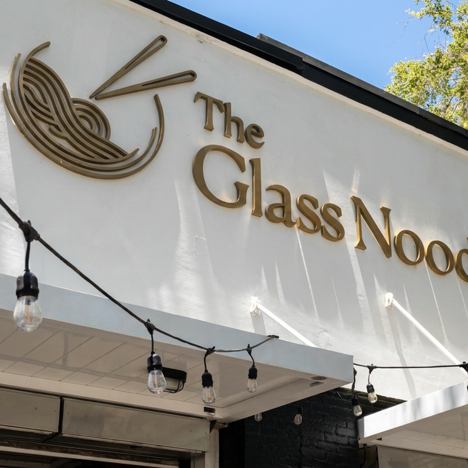 White restaurant facade displaying The Glass Noodle sign and noodle bowl logo beneath string lights and a leafy tree.