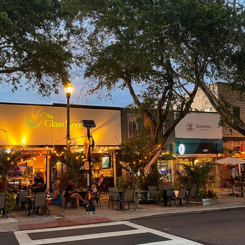 The Glass Noodle restaurant glows at dusk, with outdoor dining, string lights, trees, and neighboring Sorrento Sweets storefront.