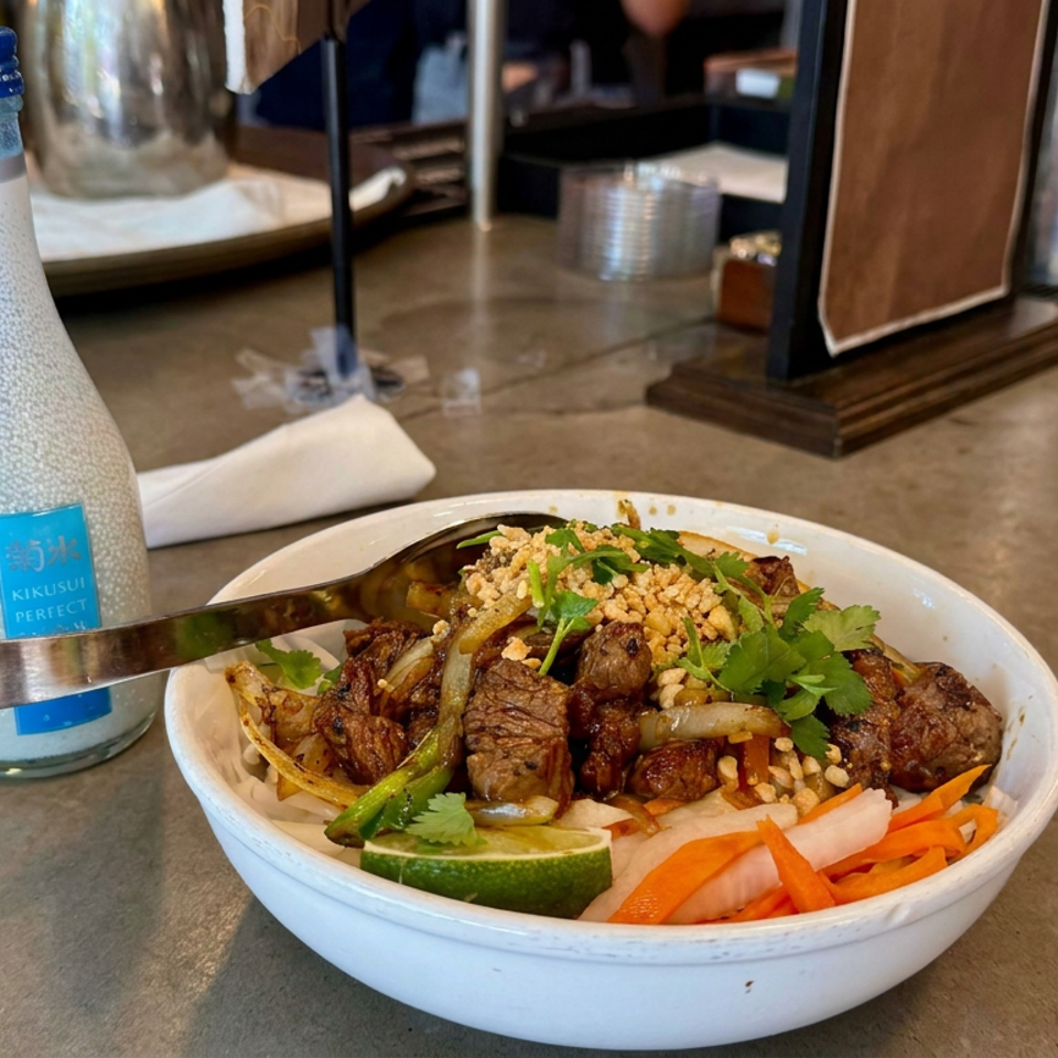 Vietnamese-style rice bowl topped with grilled beef, onions, herbs, peanuts, lime, and pickled vegetables beside a bottle of sake.