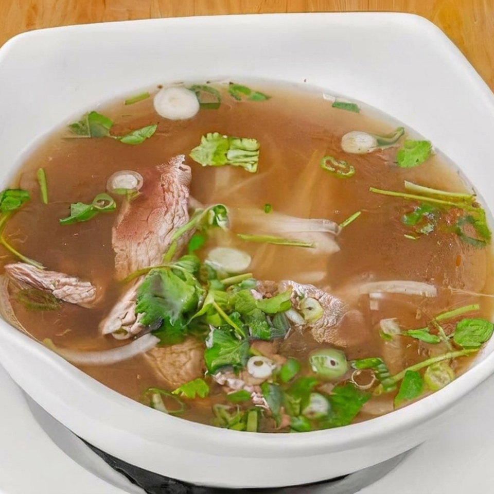 Bowl of clear beef noodle soup garnished with cilantro, green onions, and sliced jalapeño, served with lime wedges.